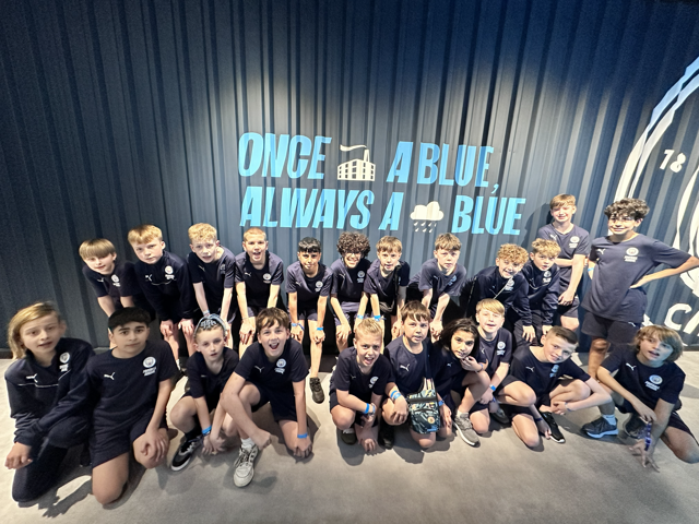 Group of children in navy sports attire posing in front of a sign with the text 'ONCE A BLUE, ALWAYS A BLUE.'