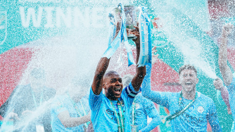 A football team celebrates winning a trophy with confetti and champagne. Players are in light blue kits.