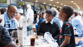 A group of people laughing and dining at a table in a busy indoor venue. They appear to be enjoying their time, surrounded by glasses, bottles, and a scarf. The environment is lively and social.