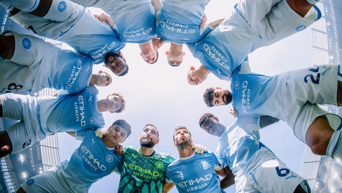 Manchester City players in a team huddle on the field, wearing light blue kits with Etihad Airways sponsor logo, viewed from below against a clear sky.