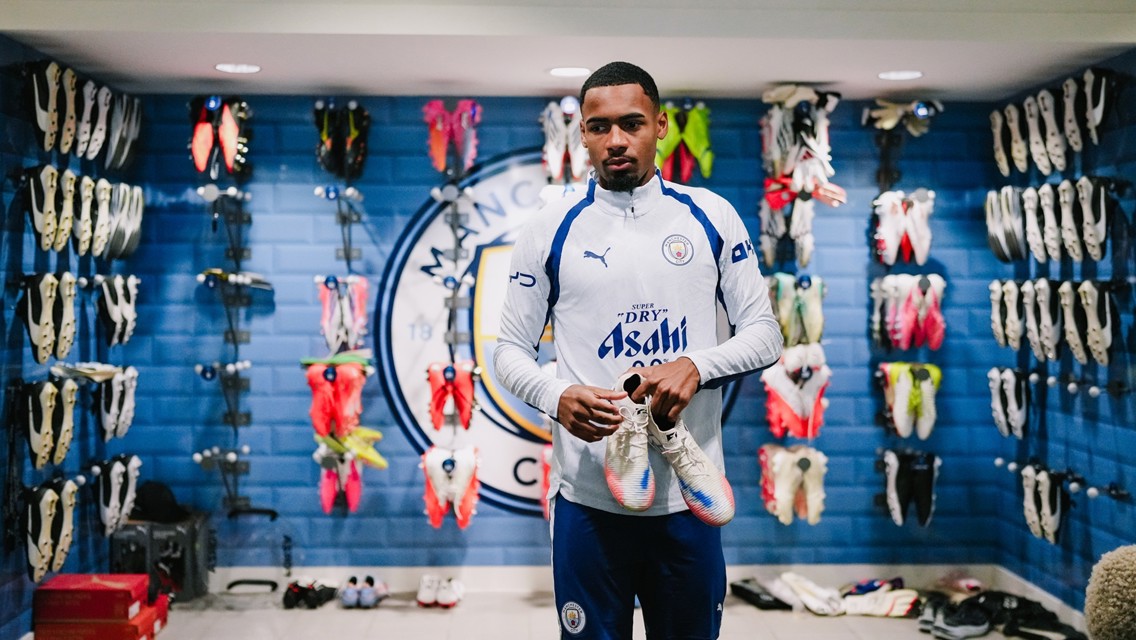 A football player holding a pair of boots in a Manchester City dressing room with boots displayed on the walls and the Manchester City logo visible.
