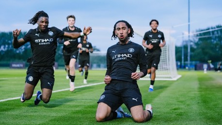 Football players wearing dark Manchester City kits celebrating a goal on a field, with two in the foreground kneeling and sliding.