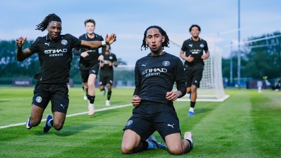 Football players wearing dark Manchester City kits celebrating a goal on a field, with two in the foreground kneeling and sliding.