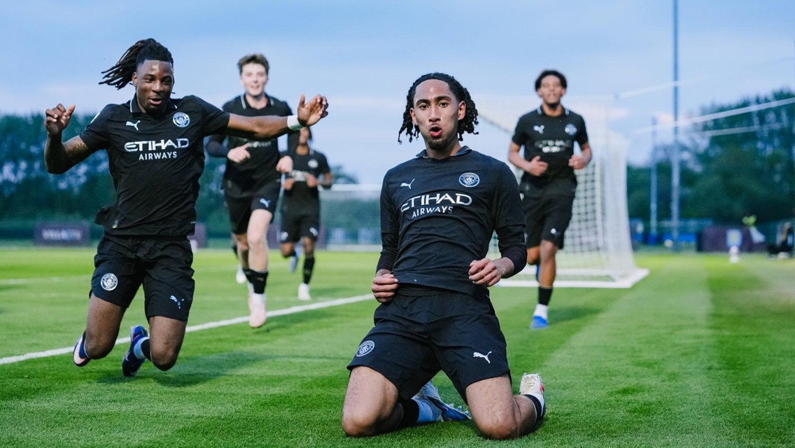 Football players wearing dark Manchester City kits celebrating a goal on a field, with two in the foreground kneeling and sliding.