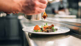 A chef drizzling sauce onto a plated dish in a professional kitchen setting, featuring meat, vegetables, and garnishes.