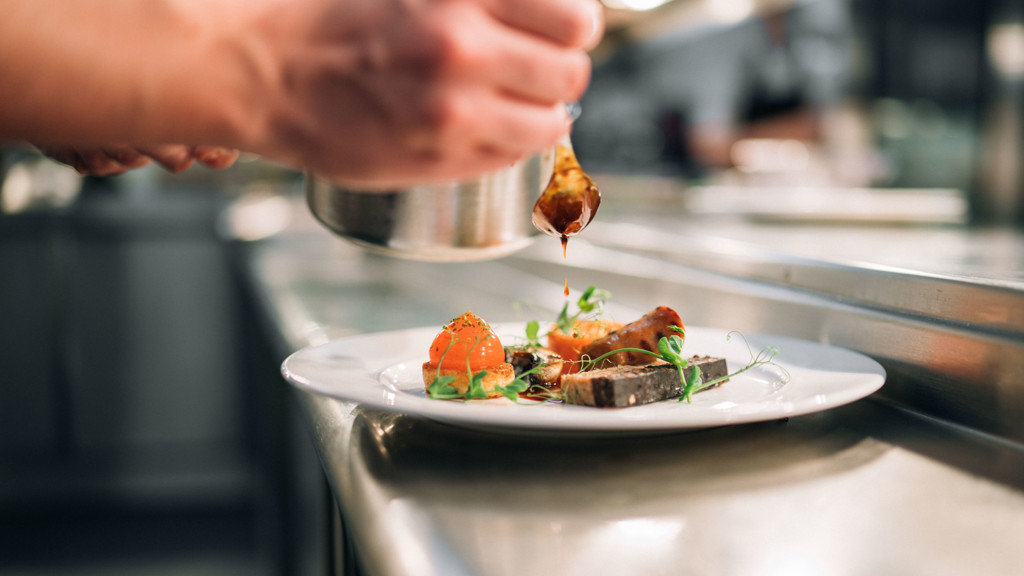 A chef drizzling sauce onto a plated dish in a professional kitchen setting, featuring meat, vegetables, and garnishes.