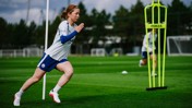 A female footballer wearing a Manchester City training kit is sprinting on a grass field. Training equipment and goalposts are visible in the background.