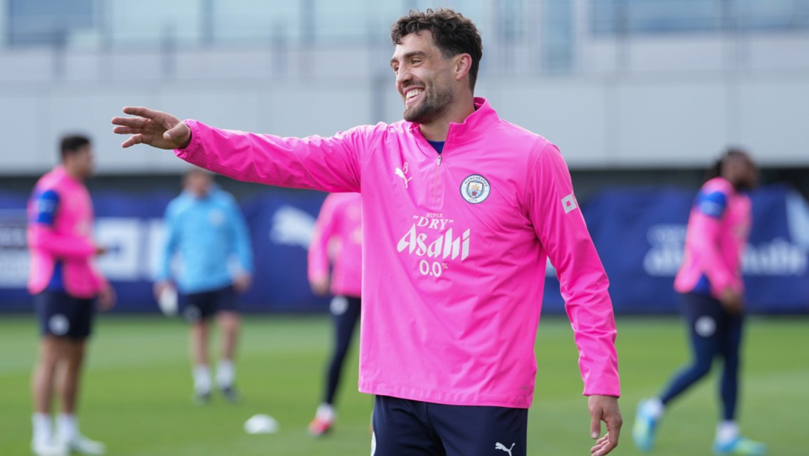 A Man City player wearing a pink training jacket gestures during a training session, with other players and coaches visible in the background.