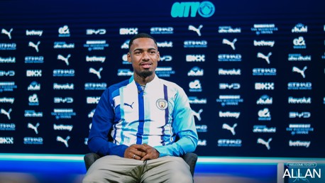 A person sits in front of a Manchester City backdrop wearing a team jacket, with various sponsor logos displayed behind. The text 'Allan' is featured prominently at the bottom right.