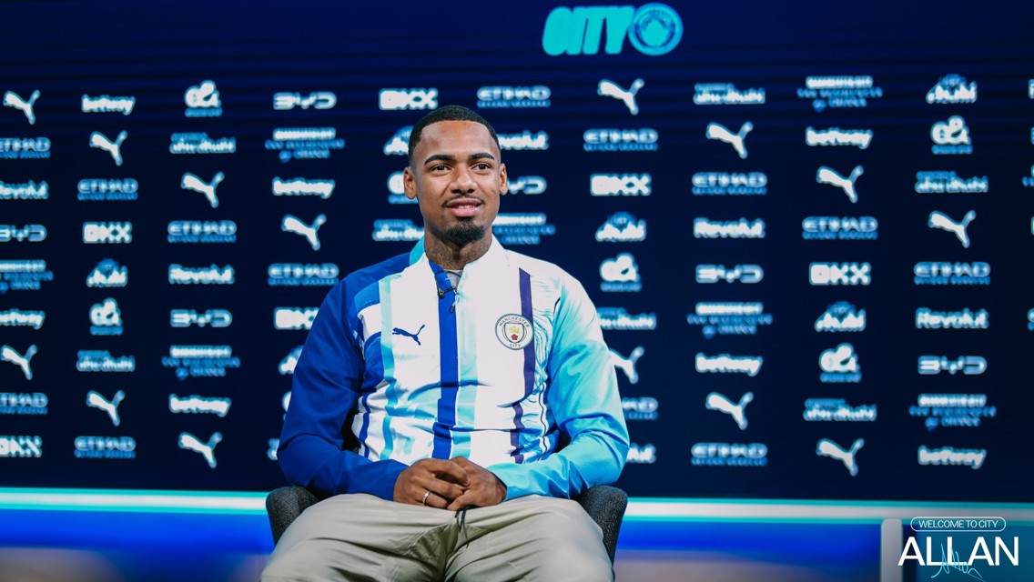 A person sits in front of a Manchester City backdrop wearing a team jacket, with various sponsor logos displayed behind. The text 'Allan' is featured prominently at the bottom right.