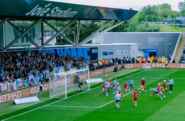 Players in light blue and red jerseys compete near the goal at Joie Stadium with a crowd of spectators.