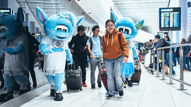 Manchester City mascots Moonbeam and Moonchester walking through an airport with people carrying suitcases, celebrating Premier League 2022/23 victory.