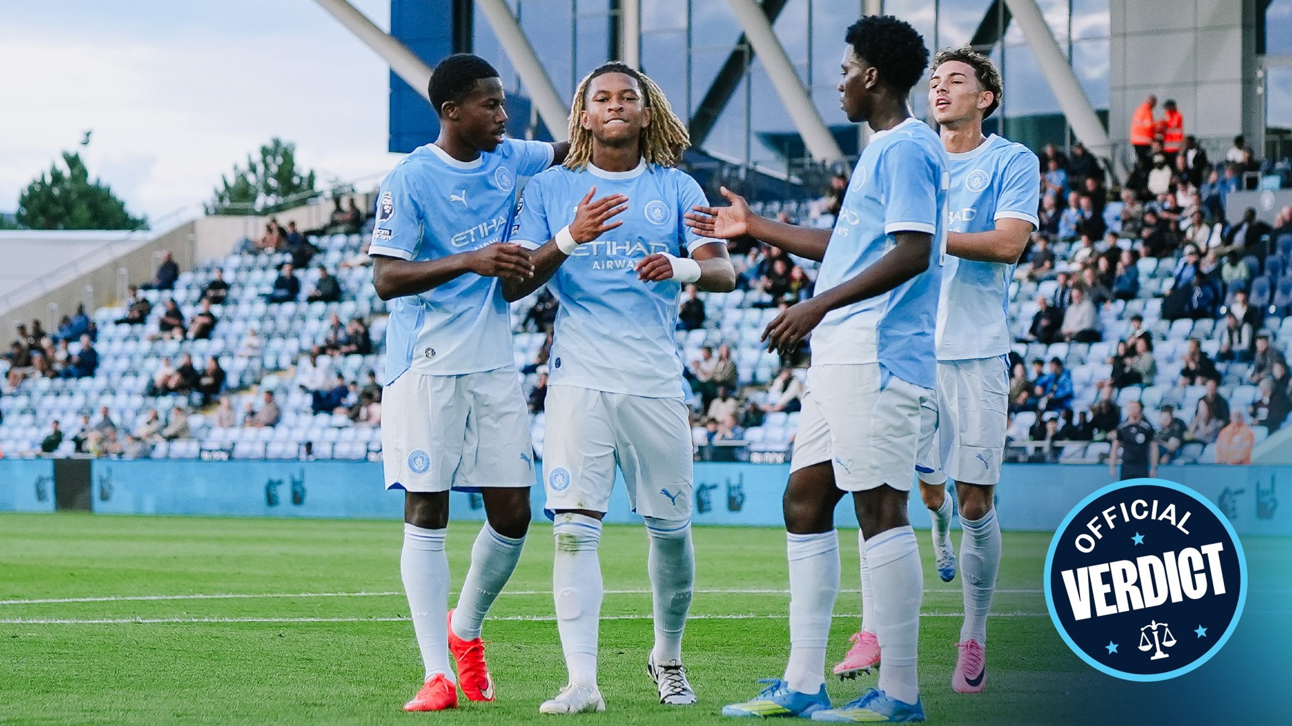 Four Manchester City players wearing light blue kits celebrate on a football field with an 'Official Verdict' badge in the corner.