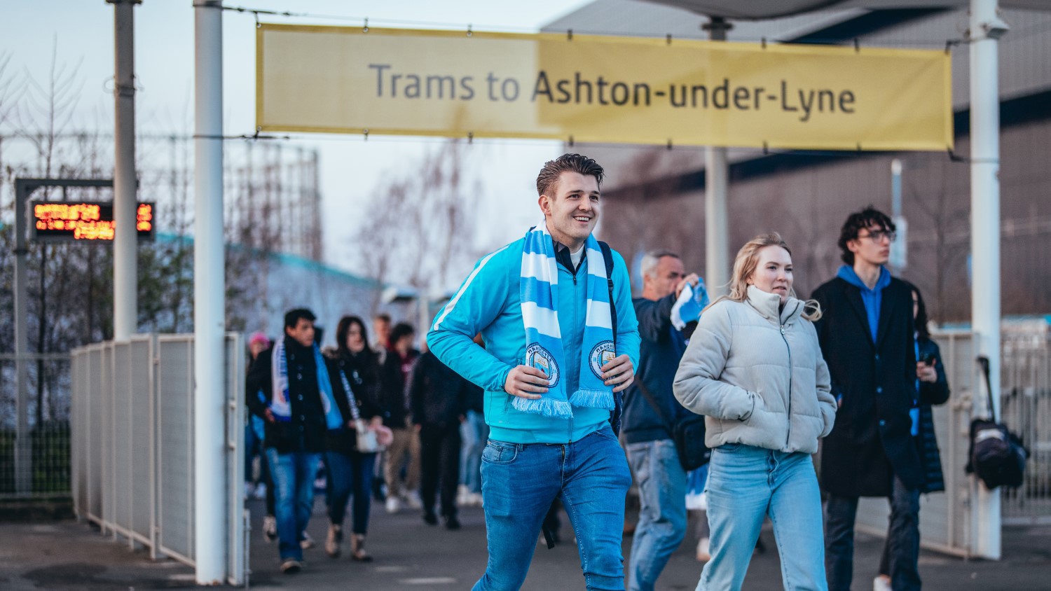 Group of people walking under a yellow sign that reads 'Trams to Ashton-under-Lyne'.