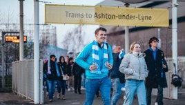 Group of people walking under a yellow sign that reads 'Trams to Ashton-under-Lyne'.