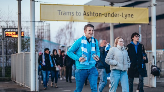 Group of people walking under a yellow sign that reads 'Trams to Ashton-under-Lyne'.
