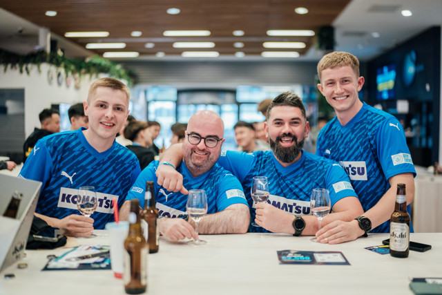 A group of Manchester City fans wearing blue Puma jerseys with the Komatsu logo, celebrating in a pub with drinks on the table.