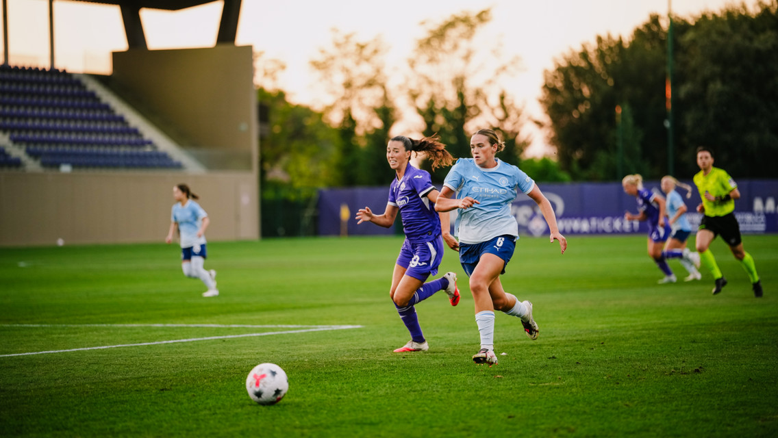 Two women soccer players competing for the ball during a match on a grass field with a referee in the background.