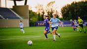 Two women soccer players competing for the ball during a match on a grass field with a referee in the background.