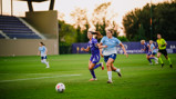 Two women soccer players competing for the ball during a match on a grass field with a referee in the background.