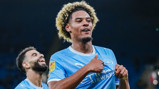 A Manchester City player celebrating a goal on the field wearing a light blue jersey. Another player is visible in the background.
