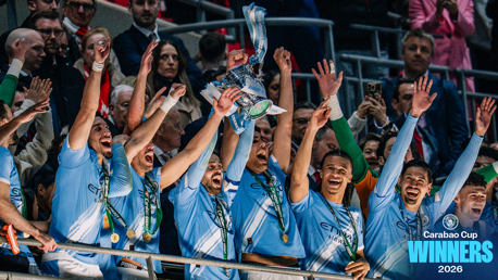 Manchester City players celebrating a Carabao Cup victory in 2026, holding the trophy with medals around their necks. The team is wearing light blue jerseys in front of a cheering crowd.