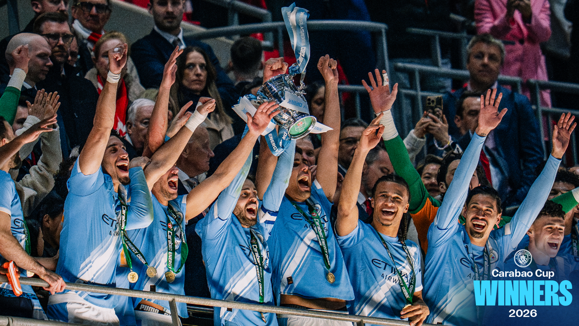 Manchester City players celebrating a Carabao Cup victory in 2026, holding the trophy with medals around their necks. The team is wearing light blue jerseys in front of a cheering crowd.