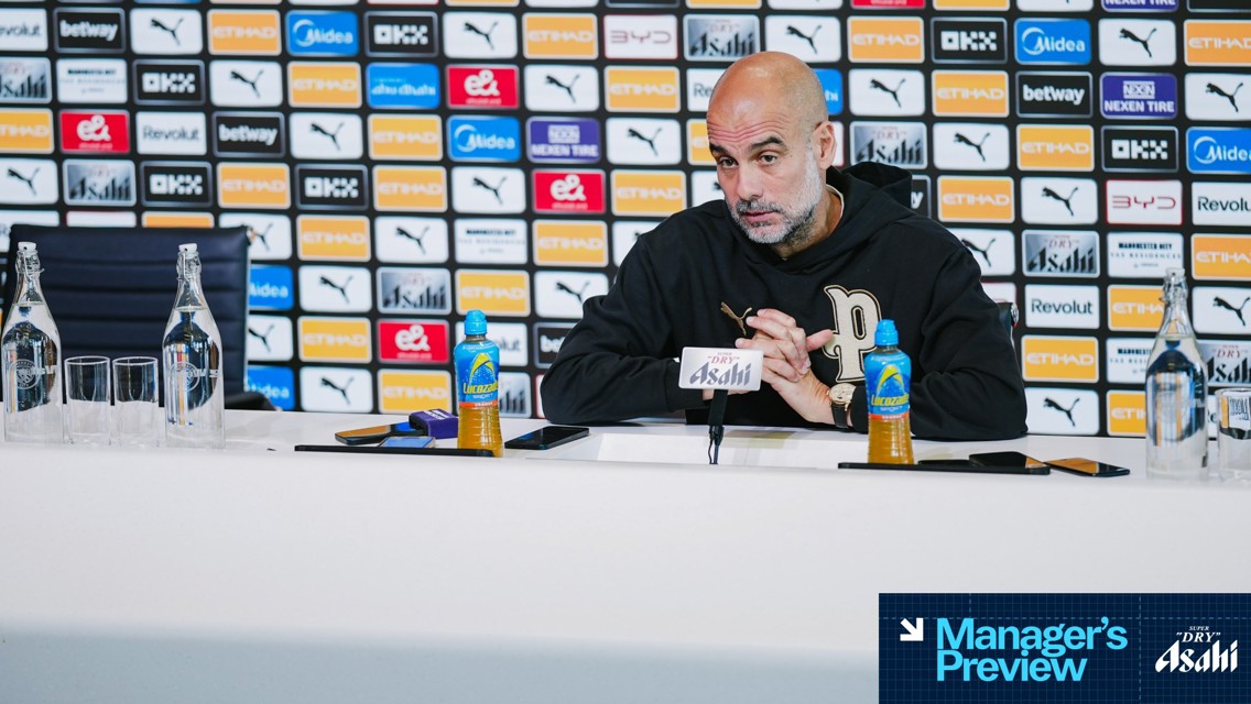 A press conference at a stadium with a seated person behind microphones. There are logos on the backdrop and branded bottles on the table.
