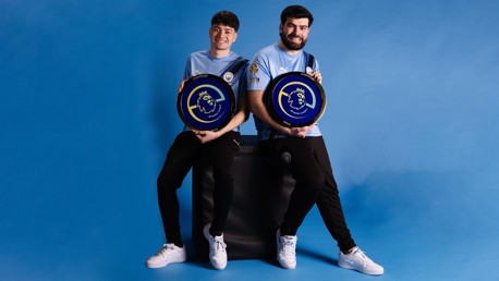 Two individuals in Manchester City jerseys holding ePremier League champion plaques against a blue background.