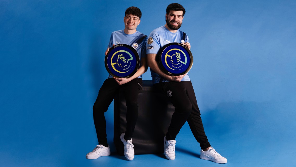 Two individuals in Manchester City jerseys holding ePremier League champion plaques against a blue background.