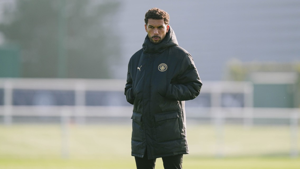 A person wearing a black jacket with Manchester City branding stands outdoors on a sports field.