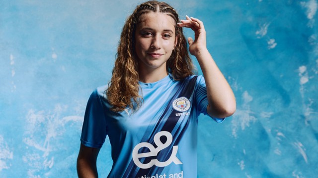 A woman wearing a Man City Esport jersey with the Manchester City crest on it, standing against a blue background.