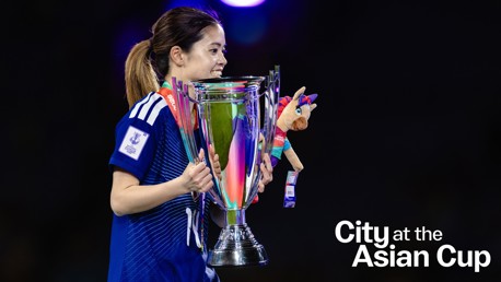 A person in a blue sports jersey holding the Asian Cup trophy, adorned with colorful ribbons and a plush mascot.