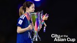 A person in a blue sports jersey holding the Asian Cup trophy, adorned with colorful ribbons and a plush mascot.