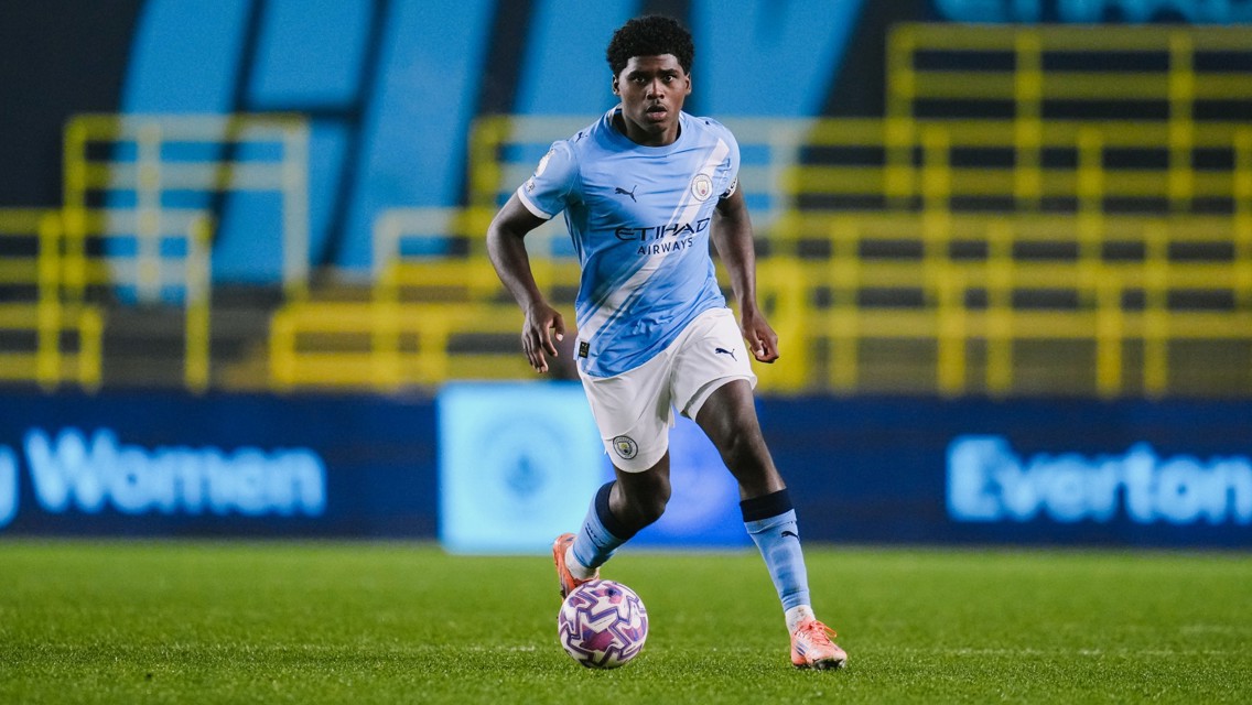 A footballer in a Manchester City uniform dribbling a ball on the field with stands in the background.