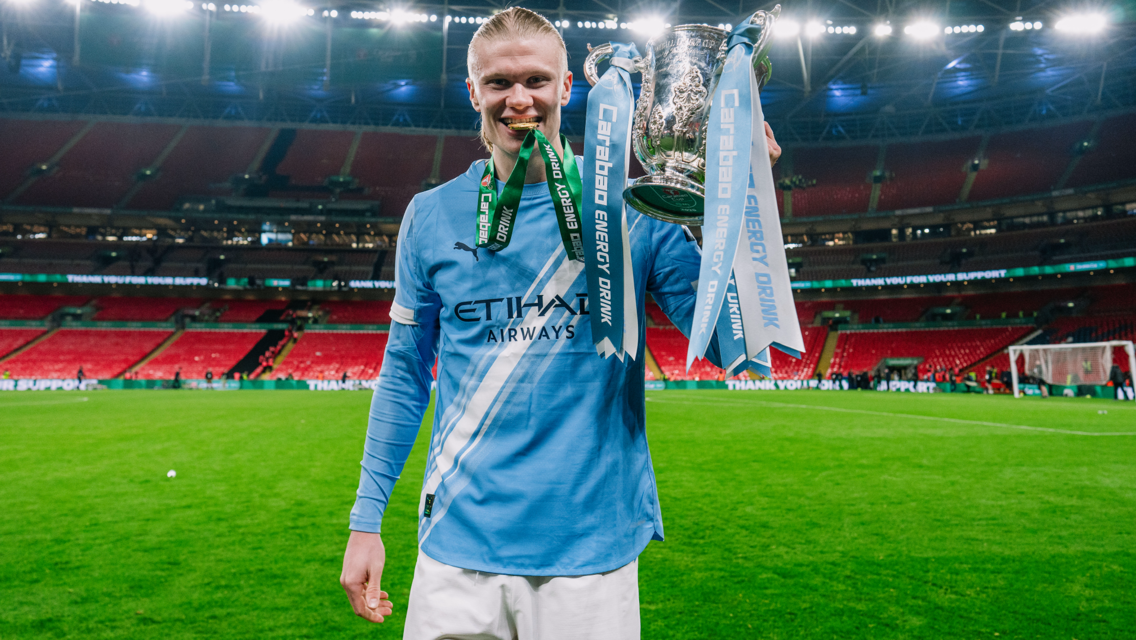 A football player in a Manchester City kit holding the Carabao Cup on a football pitch.