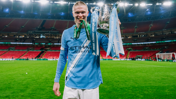 A football player in a Manchester City kit holding the Carabao Cup on a football pitch.