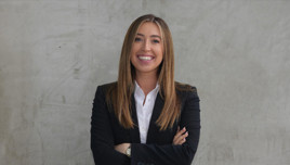 A woman in a business suit stands with her arms crossed in front of a concrete wall.