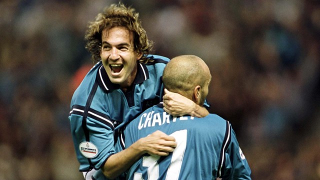 Two soccer players celebrating a goal on the field, wearing blue jerseys. One player is hugging the other, showing excitement and joy.