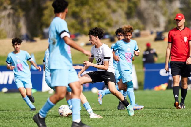 Kids playing a soccer match on a grass field. The focus is on a player wearing a white shirt and black shorts surrounded by opponents in light blue kits. A referee in red is visible in the background.