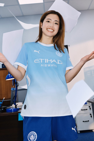 A person wearing a Manchester City football kit stands in an office, surrounded by floating sheets of paper.