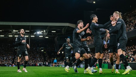 Manchester City players wearing black kits celebrate a goal during a match in a crowded stadium. The team jerseys display sponsor logos including Etihad Airways.