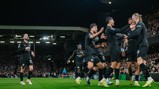 Manchester City players wearing black kits celebrate a goal during a match in a crowded stadium. The team jerseys display sponsor logos including Etihad Airways.