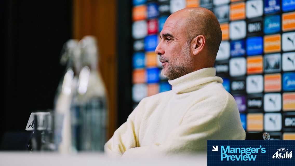An individual in a white turtleneck sweater sits at a press conference table with a blurred face. The background features a colorful display of logos.