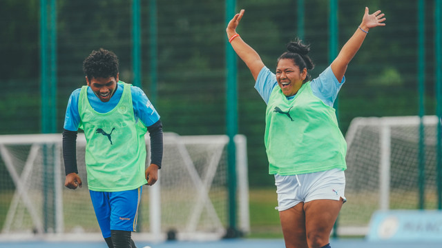 Two football players in blue shorts and green bibs celebrating on the field, with one raising arms and the other preparing to celebrate.