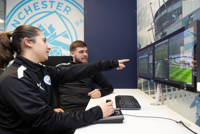 Two people in Man City gear monitor multiple screens showing sports fields. The Manchester City logo is visible in the background.
