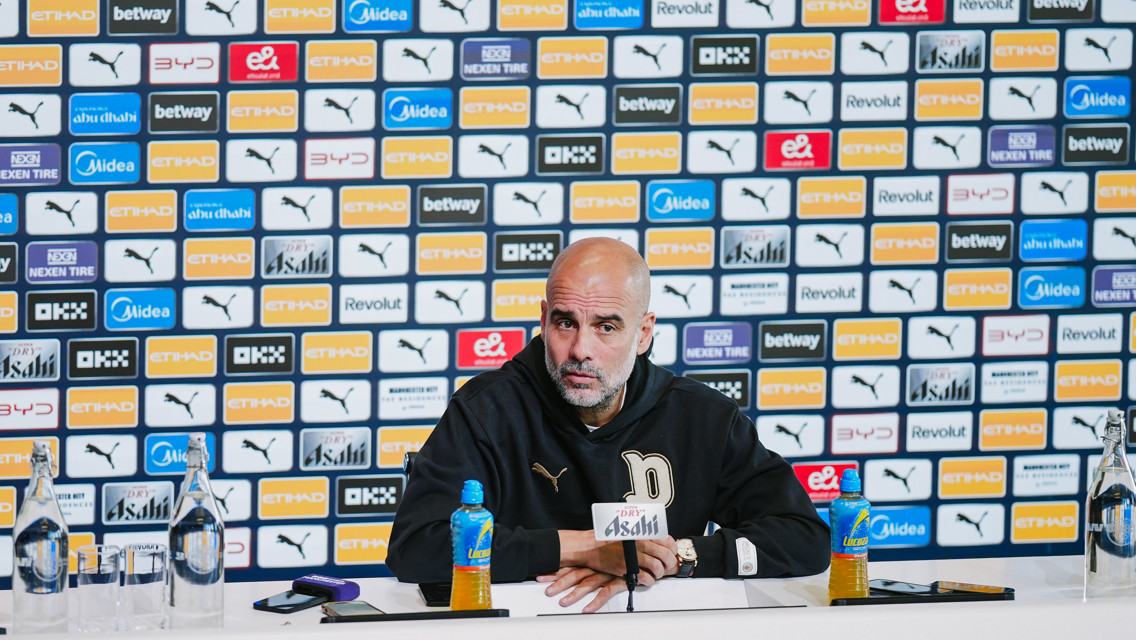 A person at a press conference table wearing a black jacket with the letter 'P' logo. Background shows a wall with logos including Puma and Etihad.