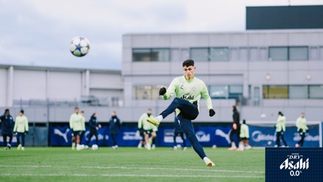 A Manchester City player in training gear kicks a football on a training field with a Champions League ball in view. Asahi 0.0 branding is in the bottom right corner.