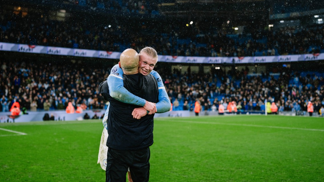 Two people hugging on a football field in front of a cheering crowd, one wearing a Manchester City kit.