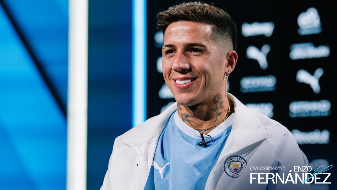 A person wearing a Manchester City jacket is smiling in front of a background with Puma and Manchester City logos. The text 'Welcome to City Enzo Fernandez' is visible.
